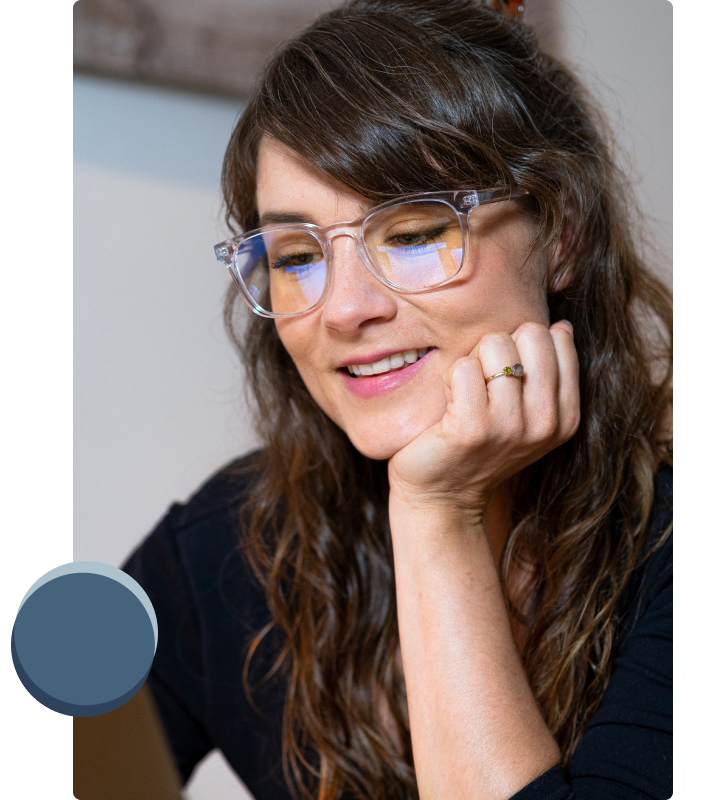 Kvinna med lockigt hår och lugg sitter framför sin datorn med Luxreaders blue light skärmglasögon och ett leende på läpparna
